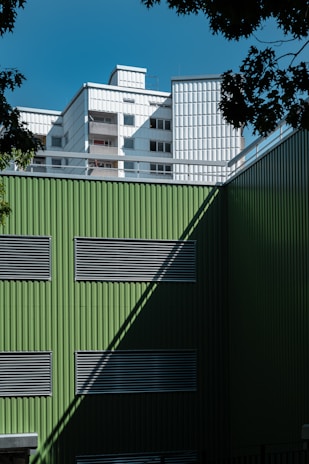 A modern architectural scene featuring a white, multi-story building with grid-like glass panels. In the foreground, there is a green building with corrugated metal cladding and horizontal vents. The sky is clear blue, and the upper part of a tree with dark green leaves frames the scene.