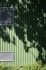 A green metal wall with vertical ridges features two rectangular ventilators. Shadows of leaves create patterns on the surface. At the bottom, a row of small green plants adds a natural element.