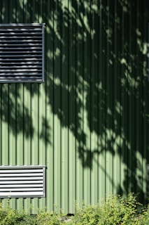 A green metal wall with vertical ridges features two rectangular ventilators. Shadows of leaves create patterns on the surface. At the bottom, a row of small green plants adds a natural element.