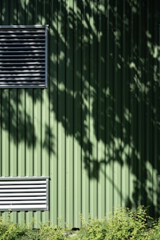 A green metal wall with vertical ridges features two rectangular ventilators. Shadows of leaves create patterns on the surface. At the bottom, a row of small green plants adds a natural element.