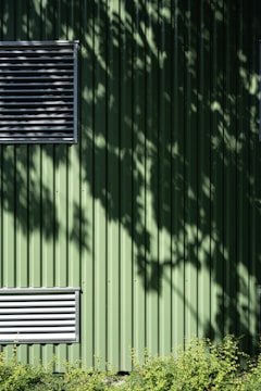 A green metal wall with vertical ridges features two rectangular ventilators. Shadows of leaves create patterns on the surface. At the bottom, a row of small green plants adds a natural element.