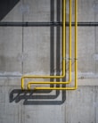 A concrete wall with three vertical yellow pipes running parallel, casting long shadows. The wall has some visible water stains and a rough texture, showcasing industrial design elements.