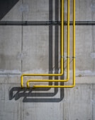 A concrete wall with three vertical yellow pipes running parallel, casting long shadows. The wall has some visible water stains and a rough texture, showcasing industrial design elements.