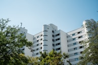 A modern, multi-story apartment building with a white façade and cylindrical features on the roof. The building is surrounded by lush green trees, and the sky above is clear and blue.