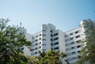 A modern, multi-story apartment building with a white façade and cylindrical features on the roof. The building is surrounded by lush green trees, and the sky above is clear and blue.