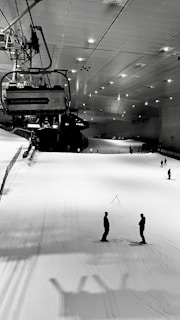 An indoor ski slope with ski lifts overhead, illuminated by ceiling lights. Several people are skiing on the white snowy surface, which is bordered by safety nets on the left side. The setting is serene and industrial in an enclosed environment.