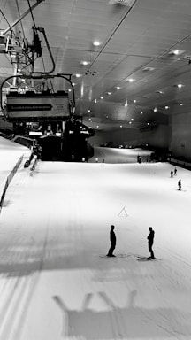 An indoor ski slope with ski lifts overhead, illuminated by ceiling lights. Several people are skiing on the white snowy surface, which is bordered by safety nets on the left side. The setting is serene and industrial in an enclosed environment.