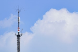 A tall communication tower extends into the sky, surrounded by clear blue skies and a few fluffy white clouds. The tower is equipped with various antennas and equipment.
