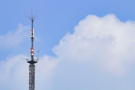 A tall communication tower extends into the sky, surrounded by clear blue skies and a few fluffy white clouds. The tower is equipped with various antennas and equipment.