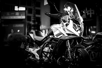 A sleek motorcycle parked in a dimly lit environment with prominent reflective surfaces and angular design features. The background has blurred signage and a muted street scene, suggesting an urban setting at night.