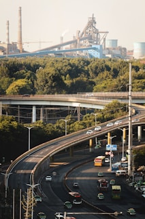 An elevated highway snakes through an urban landscape, with multiple vehicles including cars and buses traveling along it. In the background, an industrial area with large factories and smokestacks is visible. Dense greenery surrounds the highway, providing a natural contrast to the industrial structures.