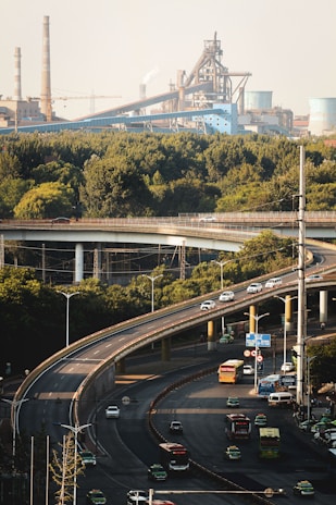 An elevated highway snakes through an urban landscape, with multiple vehicles including cars and buses traveling along it. In the background, an industrial area with large factories and smokestacks is visible. Dense greenery surrounds the highway, providing a natural contrast to the industrial structures.