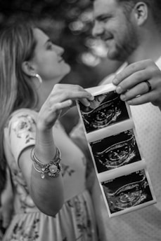 A black and white image of a couple holding sonogram pictures. The woman, wearing a floral dress and bracelets, lovingly gazes at her partner, who smiles back. The focus is on the sonogram images being held by the couple, capturing a sense of joy and anticipation.