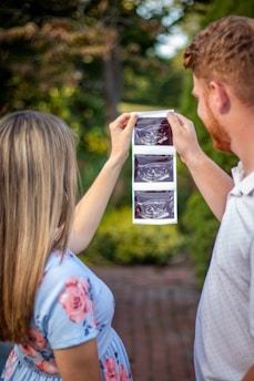A couple is holding up ultrasound images in an outdoor setting, surrounded by greenery. The woman is wearing a light blue dress with pink floral patterns, and the man is in a light-colored polo shirt.