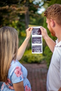 A couple is holding up ultrasound images in an outdoor setting, surrounded by greenery. The woman is wearing a light blue dress with pink floral patterns, and the man is in a light-colored polo shirt.