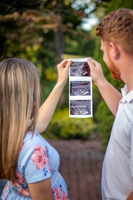 A couple is holding up ultrasound images in an outdoor setting, surrounded by greenery. The woman is wearing a light blue dress with pink floral patterns, and the man is in a light-colored polo shirt.