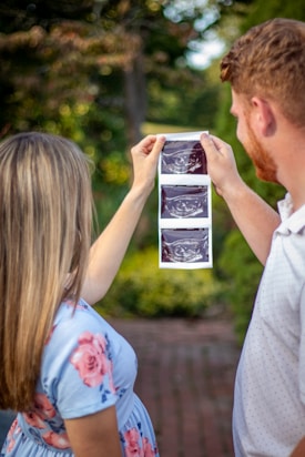 A couple is holding up ultrasound images in an outdoor setting, surrounded by greenery. The woman is wearing a light blue dress with pink floral patterns, and the man is in a light-colored polo shirt.