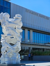 A large abstract white sculpture stands prominently in front of a modern building with the sign 'İstanbul Modern'. The building features large glass windows, and there are a few people casually standing nearby, enjoying the sunny day.