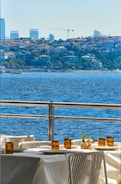 A scenic view of a waterfront with a dining table and chairs arranged outdoors. The table is set with white tablecloths, amber glass candles, and condiment holders. In the background, a sprawling urban landscape is visible with buildings and a crane, while the foreground features a vibrant blue body of water.