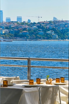 A scenic view of a waterfront with a dining table and chairs arranged outdoors. The table is set with white tablecloths, amber glass candles, and condiment holders. In the background, a sprawling urban landscape is visible with buildings and a crane, while the foreground features a vibrant blue body of water.