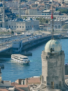 A bustling cityscape featuring a historic mosque with multiple domes and minarets, a busy bridge with traffic, a large white ferry navigating the waterway, and a prominent tower in the foreground adorned with a flag.