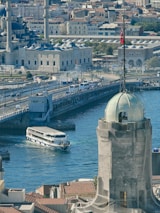 A bustling cityscape featuring a historic mosque with multiple domes and minarets, a busy bridge with traffic, a large white ferry navigating the waterway, and a prominent tower in the foreground adorned with a flag.