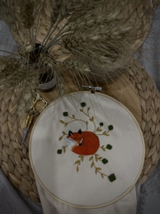 An embroidery hoop displays a detailed image of a curled-up fox surrounded by leaves. The background features a woven mat and a small pair of golden embroidery scissors. A bundle of dried wheat stalks is placed nearby, adding a natural, rustic feel.