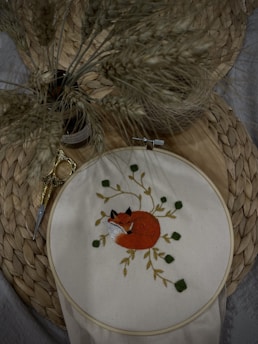 An embroidery hoop displays a detailed image of a curled-up fox surrounded by leaves. The background features a woven mat and a small pair of golden embroidery scissors. A bundle of dried wheat stalks is placed nearby, adding a natural, rustic feel.