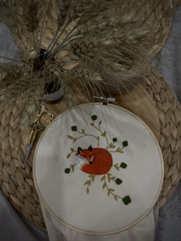 An embroidery hoop displays a detailed image of a curled-up fox surrounded by leaves. The background features a woven mat and a small pair of golden embroidery scissors. A bundle of dried wheat stalks is placed nearby, adding a natural, rustic feel.