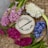 An embroidery hoop with the words 'embroidery by Liza' in black thread is positioned on a wooden surface. Surrounding the hoop are vibrant flowers in shades of pink, white, red, and blue placed on top of woven straw or wicker.
