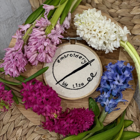 An embroidery hoop with the words 'embroidery by Liza' in black thread is positioned on a wooden surface. Surrounding the hoop are vibrant flowers in shades of pink, white, red, and blue placed on top of woven straw or wicker.