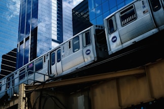 A train of the Chicago Transit Authority (CTA) is moving on an elevated track through a cityscape of tall, reflective skyscrapers. The buildings feature glass facades that mirror the sky and surrounding structures.