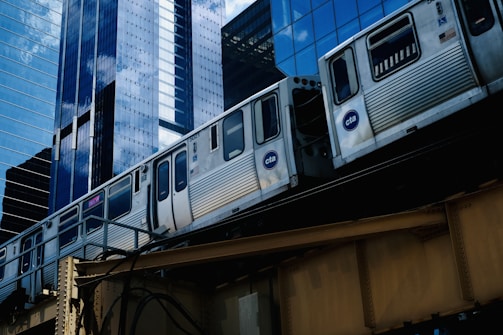 A train of the Chicago Transit Authority (CTA) is moving on an elevated track through a cityscape of tall, reflective skyscrapers. The buildings feature glass facades that mirror the sky and surrounding structures.