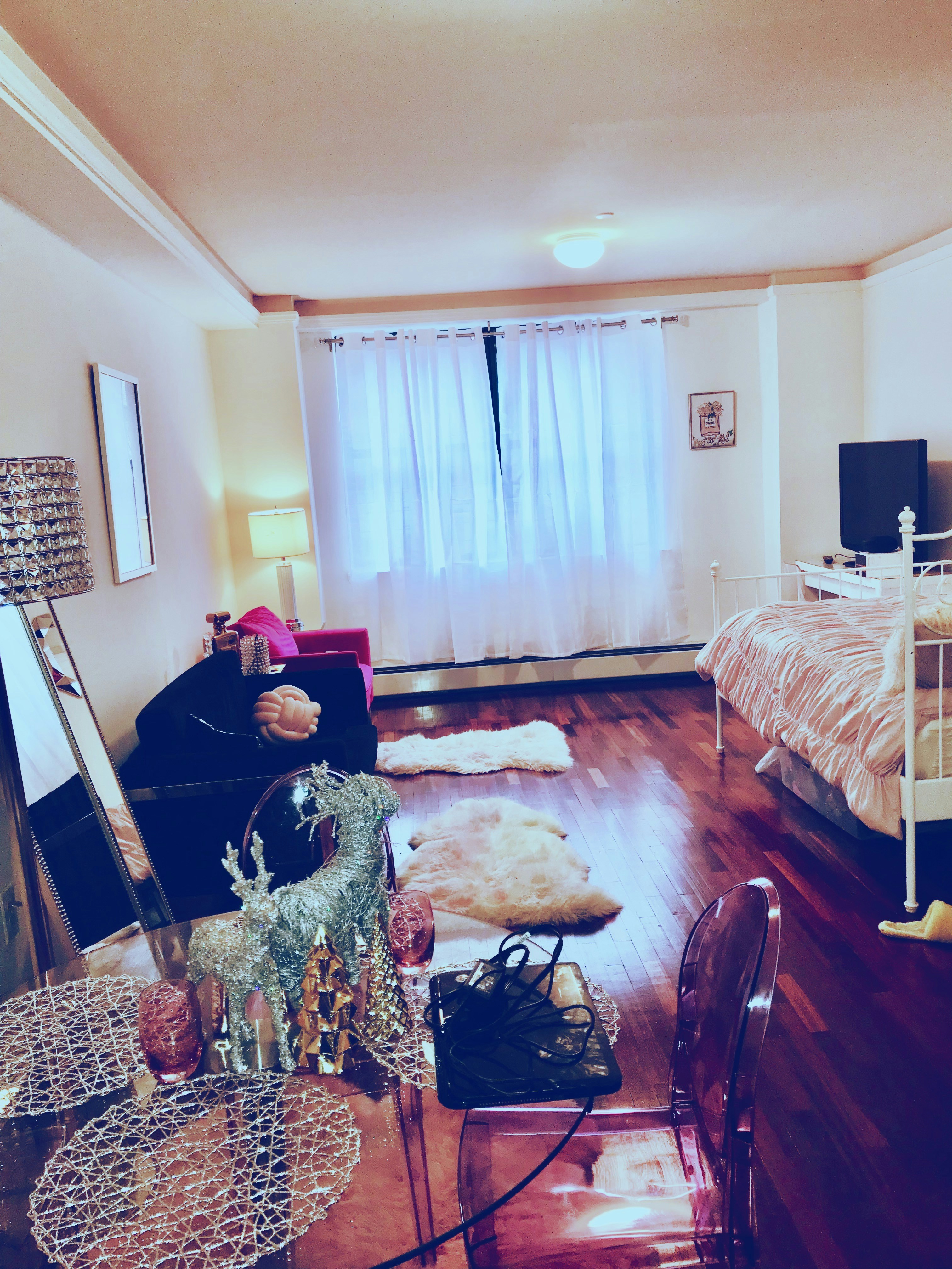 A cozy room features wooden floors, a bed with a striped comforter, a small table with decorative items like a shiny reindeer, and a clear chair. Natural light filters through sheer curtains on a large window. A black couch supports a plush pink pillow and is complemented by a nearby lamp. The walls display minimal artwork, while a TV rests on a stand against one wall.