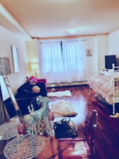 A cozy room features wooden floors, a bed with a striped comforter, a small table with decorative items like a shiny reindeer, and a clear chair. Natural light filters through sheer curtains on a large window. A black couch supports a plush pink pillow and is complemented by a nearby lamp. The walls display minimal artwork, while a TV rests on a stand against one wall.