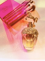 A luxurious perfume bottle with an ornate gold cap featuring a unicorn figurine is placed beside a vibrant pink container holding several makeup brushes. The setting is illuminated with a soft, warm light, casting a gentle glow over the scene.