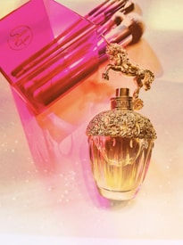 A luxurious perfume bottle with an ornate gold cap featuring a unicorn figurine is placed beside a vibrant pink container holding several makeup brushes. The setting is illuminated with a soft, warm light, casting a gentle glow over the scene.