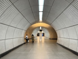 A modern underground tunnel with curved walls and a tiled ceiling, leading to a station platform. Two people are walking down the tunnel, one carrying luggage. Fluorescent lights illuminate the space, creating a futuristic ambiance.