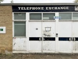 An old building with a worn and aged exterior is visible. The structure has a brick section on the left and white panels with a large black sign above displaying the words 'TELEPHONE EXCHANGE'. There are two side-by-side doors featuring metal handles and a keypad lock. A small sign reading 'Fire exit, Keep clear' is attached above the doors.