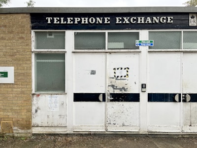 An old building with a worn and aged exterior is visible. The structure has a brick section on the left and white panels with a large black sign above displaying the words 'TELEPHONE EXCHANGE'. There are two side-by-side doors featuring metal handles and a keypad lock. A small sign reading 'Fire exit, Keep clear' is attached above the doors.