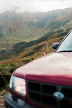 A red SUV is positioned in the foreground, partially off-frame on the right side. The background features a mountainous landscape with a winding dirt path or trail leading up the slopes. The mountains are covered in patches of green vegetation and have a rugged, rocky texture. Cloud cover is visible near the top of the mountains, creating a serene and slightly misty atmosphere.