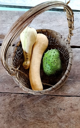 A woven basket sitting on a rustic wooden surface contains two elongated yellow squashes and a single green spiky fruit. The basket handle is curved and extends over the vegetables.