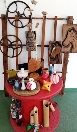 A red circular table displays various decorative vases in different colors, shapes, and sizes, surrounded by bamboo tubes and bottles. The backdrop features wooden gears and rustic wood carvings of a man and horses against a wooden lattice. Dried floral arrangements in a small vase enhance the composition.