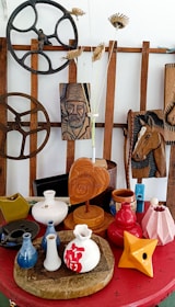 A collection of decorative items arranged on a red surface, featuring various ceramic vases, a wooden heart-shaped plaque, and intricately carved wooden panels depicting a man's face and a horse. There are also industrial-style cog wheels and dried floral arrangements.