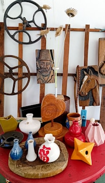 A collection of decorative items arranged on a red surface, featuring various ceramic vases, a wooden heart-shaped plaque, and intricately carved wooden panels depicting a man's face and a horse. There are also industrial-style cog wheels and dried floral arrangements.