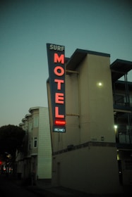 A tall, narrow sign attached to a building displays the word 'MOTEL' in bright red neon letters, with additional words 'SURF,' 'Welcome,' and 'VACANCY' visible in smaller letters. The building is located on a dimly lit street with other buildings and trees visible in the backdrop, under a dusky sky.