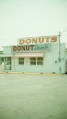 A vintage-style donut shop with a prominent sign displaying 'DONUTS' in large letters. The building has a striped red and white awning over the windows and entrance. The exterior features a light pastel color, and the foreground shows a worn parking lot surface with some yellow-painted barriers.