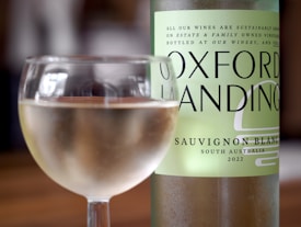 A glass of white wine is placed in front of a bottle labeled 'Oxford Landing Sauvignon Blanc South Australia 2022'. The bottle has a green label and features text emphasizing sustainable growing practices.