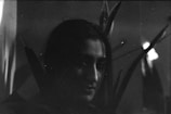 A monochrome photograph captures a person partially obscured in shadow with long hair. Tall plant leaves provide a backdrop, adding depth and texture to the composition. The low lighting creates a mysterious and moody atmosphere.