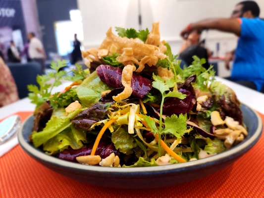 A vibrant salad featuring a variety of fresh greens, purple lettuce, and garnished with curly fried noodles and chopped nuts. The salad is served in a bowl placed on an orange table mat. In the background, blurred figures of people are visible, suggesting a cafeteria or dining setting.